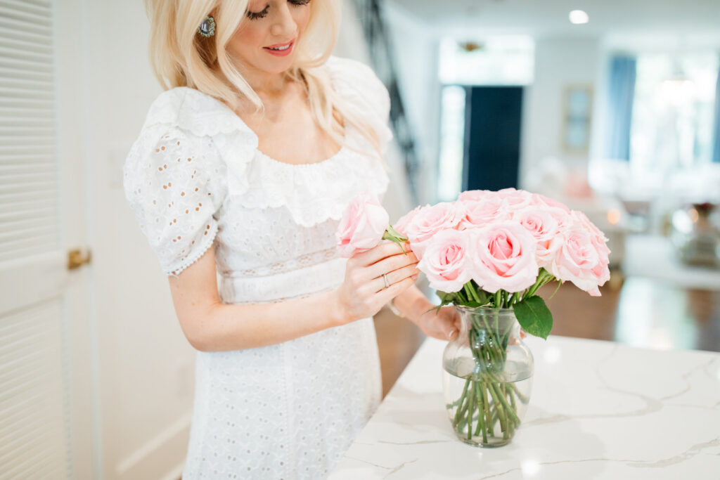 Alison Cheperdak in a white eyelet dress arranging a bouquet of pink roses in a glass vase, demonstrating grace and attention to detail in home etiquette and hosting.